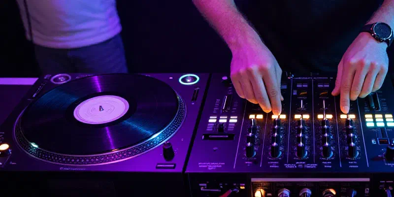 A DJ's hands on a mixer between two turntables, lit in purple and blue, with a vinyl record spinning on each deck.